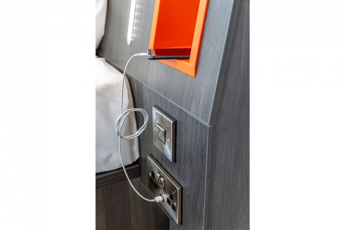 A close-up view inside an easyHotel room, featuring a dark wood-effect bedside panel with an orange alcove holding a charging smartphone. Below, UK-style power outlets and USB ports are visible.