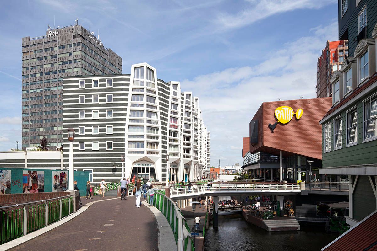 Zaandam, Netherlands: A vibrant city center with distinctive modern striped architecture, a cinema, and other buildings bordering a canal, bustling with pedestrians and cyclists on bridges and people relaxing at outdoor terraces.