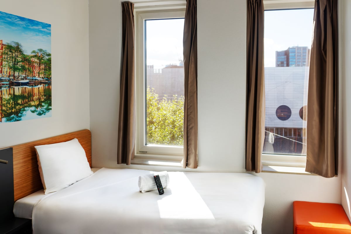 Bright easyHotel Amsterdam Arena twin room featuring a compact single bed with white linen, a wooden headboard, brown curtains, an orange stool, and a large wall art print of an Amsterdam canal scene.