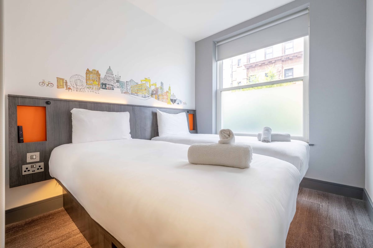 Twin room with two neatly made single beds, folded towels, and illustrated cityscape headboard.