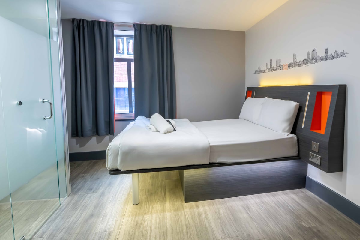 Compact hotel room with a double bed, dark curtains, and frosted glass bathroom door.