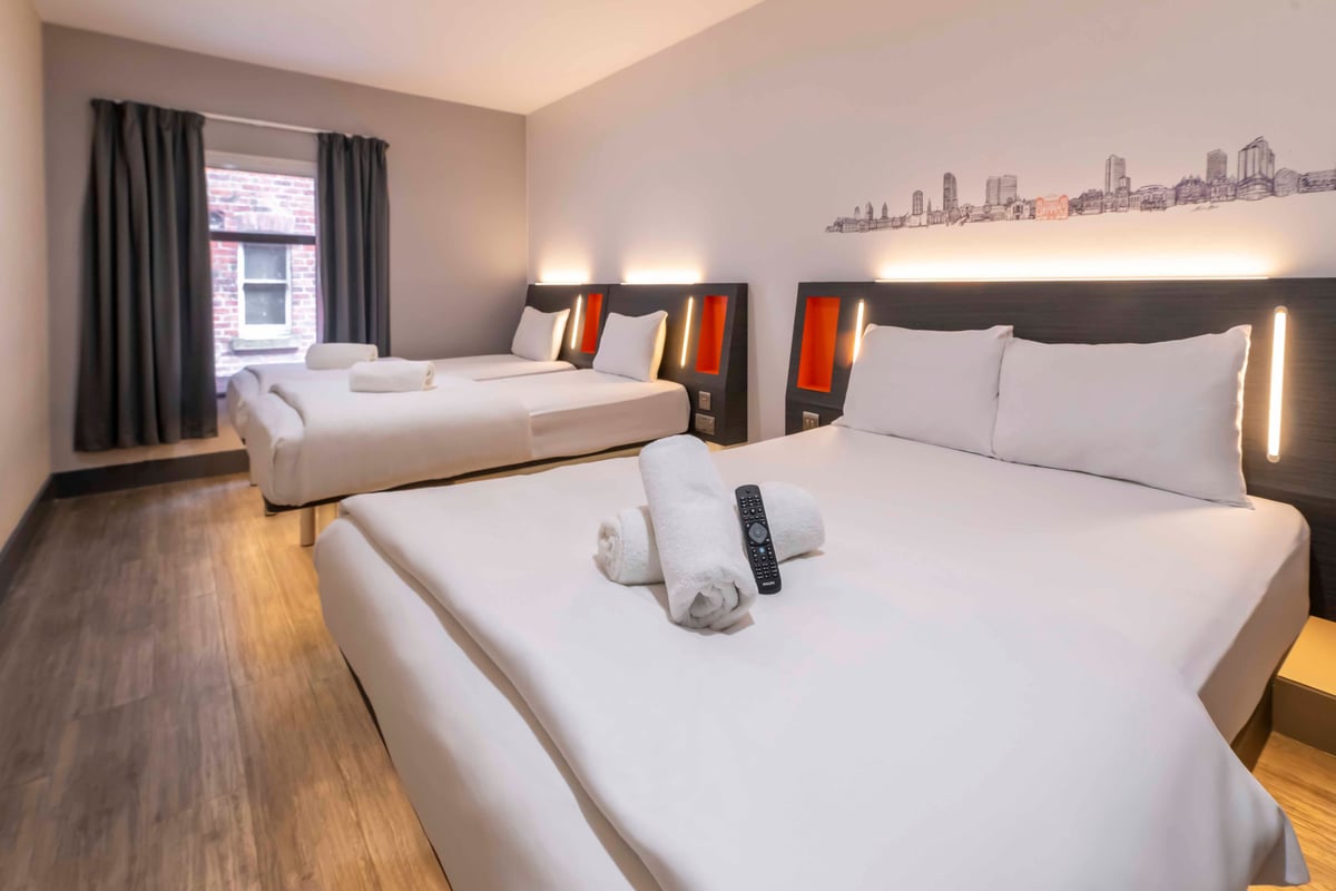 Hotel room with three beds, cityscape wall art, and towels with TV remote on the front bed.