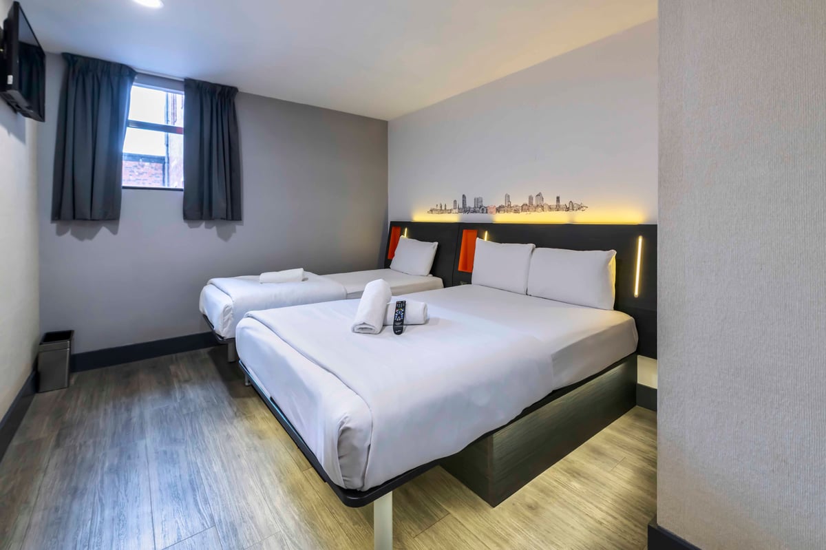 Hotel room with one double and one single bed, white linens, window and cityscape wall art.