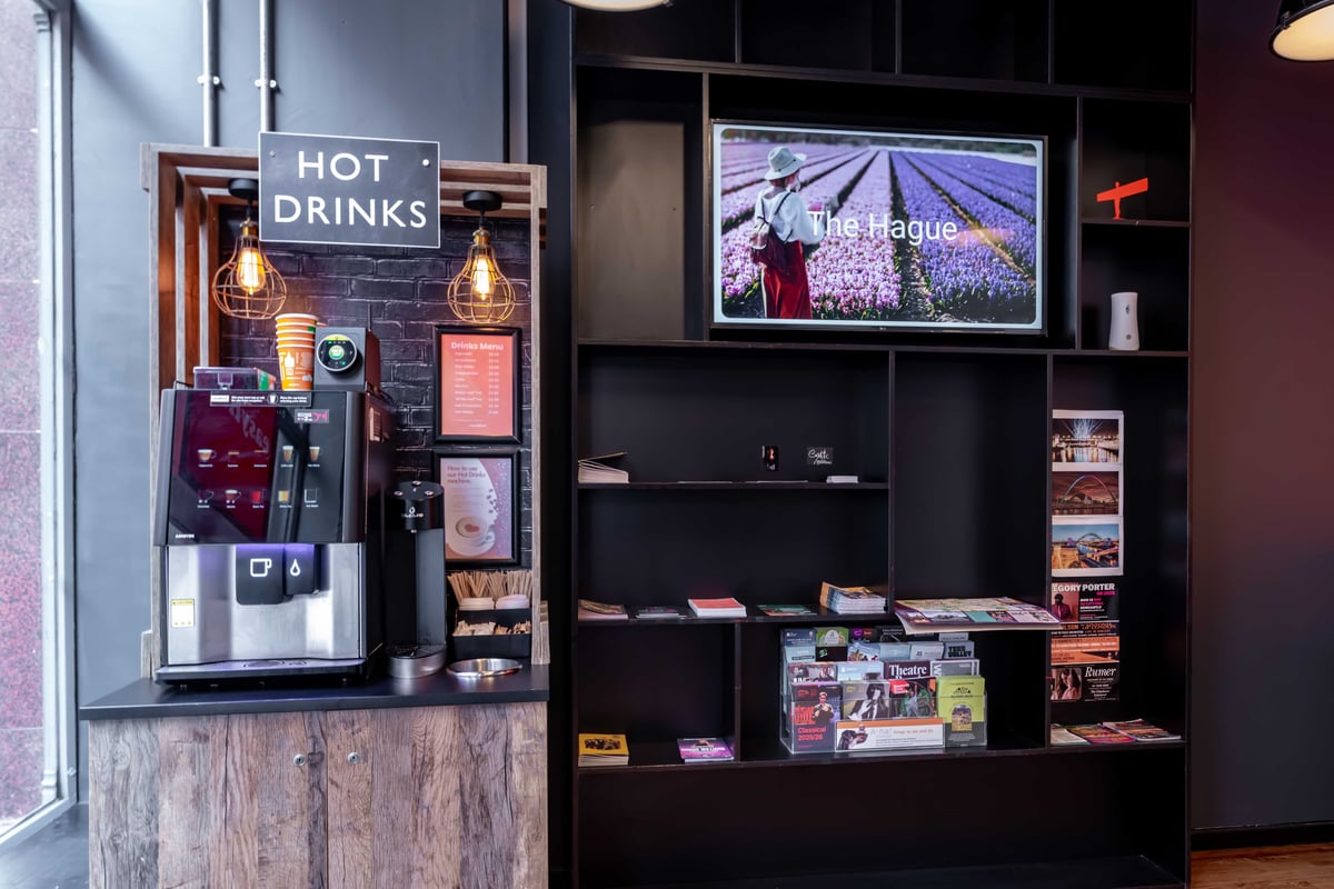 Coffee station labeled "Hot Drinks" with black shelving and screen showing.