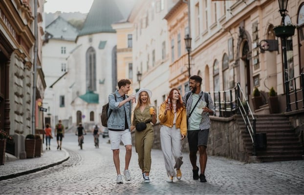 Four happy young adult friends exploring the historic cobblestone streets of Trnava, Slovakia, with old town buildings and a distinctive green-roofed church in the background.