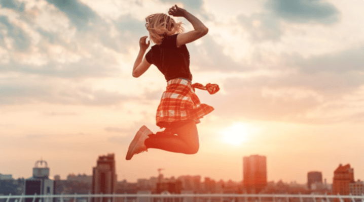 Energetic young woman jumping with joy on a city rooftop at sunset, silhouetted against a vibrant urban skyline.