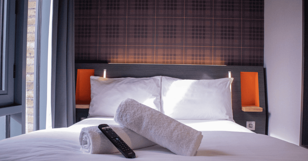 A clean, modern easyHotel room featuring a neatly made double bed with white linen, two rolled towels, and a TV remote. The room has a distinctive brown plaid wallpaper behind the headboard with orange alcove shelving, and a window revealing a brick build
