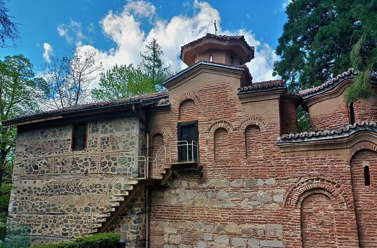Historic Boyana Church near Sofia, Bulgaria, a UNESCO World Heritage site, featuring its distinct medieval architecture with stone and red brick sections, a tiled roof, and an octagonal dome topped with a cross, under a bright blue sky.