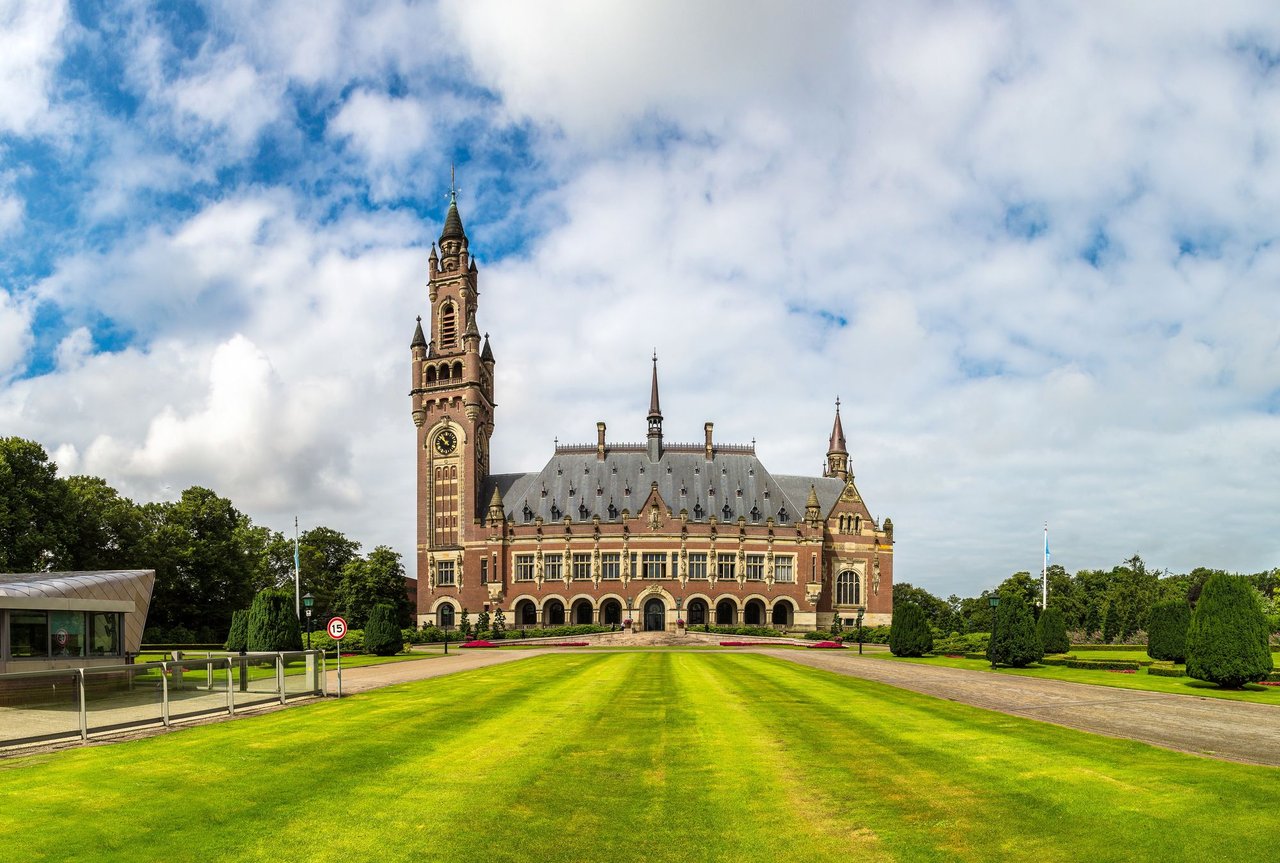 The iconic Peace Palace in The Hague, Netherlands, a grand historic brick and stone building with a prominent clock tower, surrounded by lush green lawns under a cloudy sky.