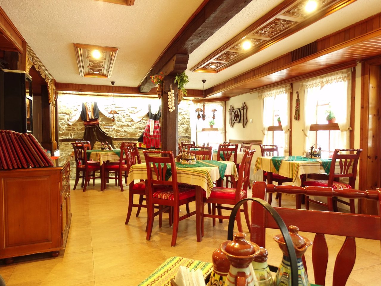 Traditional Balkan restaurant dining room with rustic wooden tables set with colorful tablecloths, red wooden chairs, exposed wooden beams, and a stone wall displaying traditional costumes.