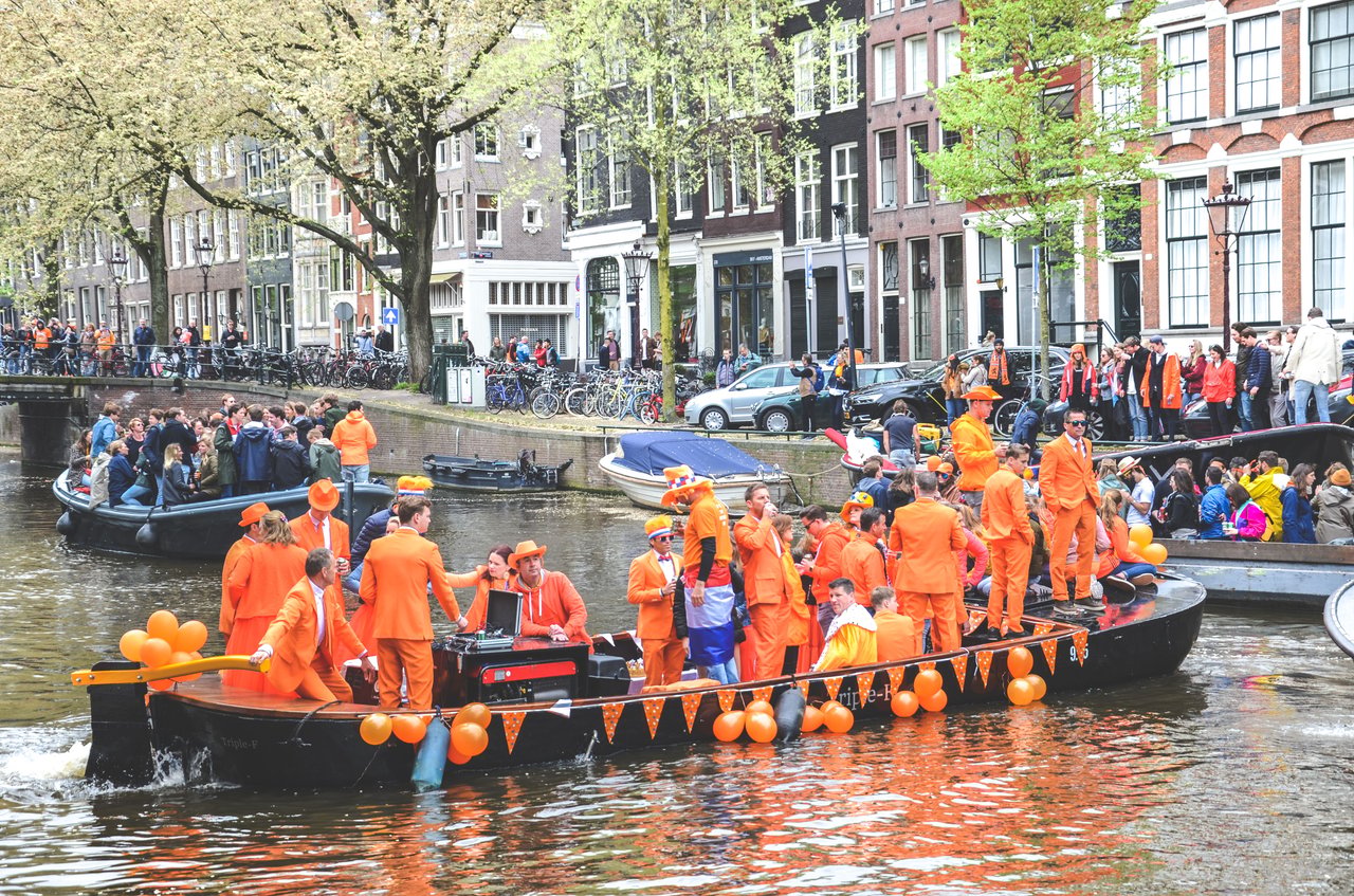 A lively King's Day celebration in Amsterdam, Netherlands, showing boats filled with people in orange attire parading along a canal, flanked by historic Dutch gabled buildings under spring trees.