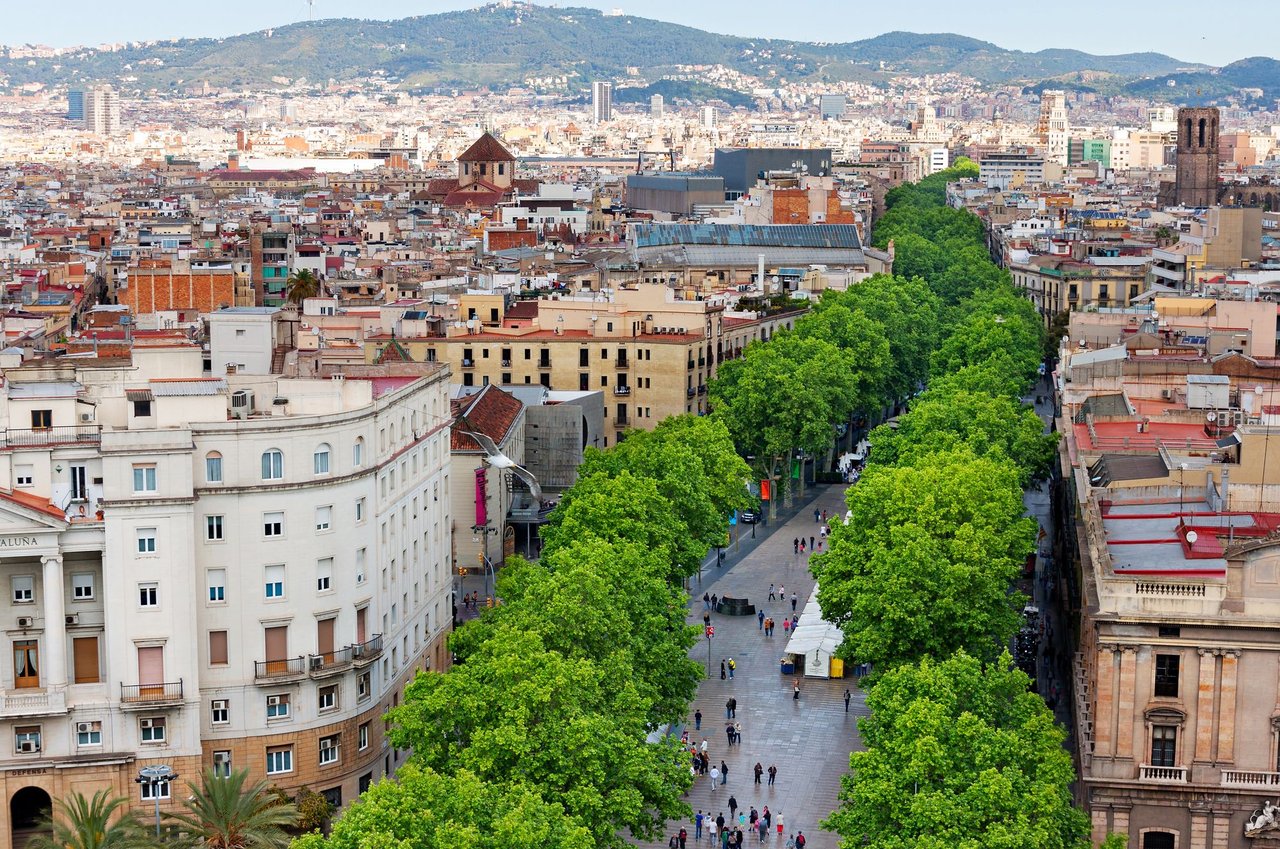 Bustling aerial view of La Rambla, Barcelona, Spain, showing the iconic tree-lined pedestrian boulevard filled with people, historic buildings, and distant mountains.