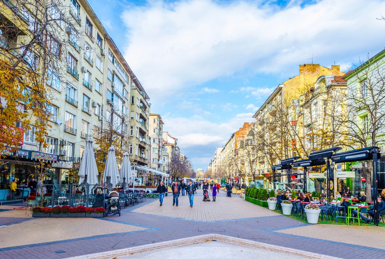 Bustling Vitosha Boulevard in Sofia, Bulgaria, a wide pedestrian street lined with historic buildings, outdoor cafes with umbrellas, and people walking on a sunny day. A distant building with a dome is visible at the end of the street.