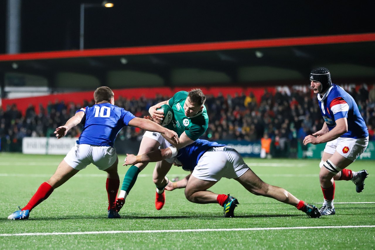 An Irish rugby player in a green jersey carrying the ball is tackled by two French players in blue jerseys during a Six Nations match under floodlights at Stade de France.