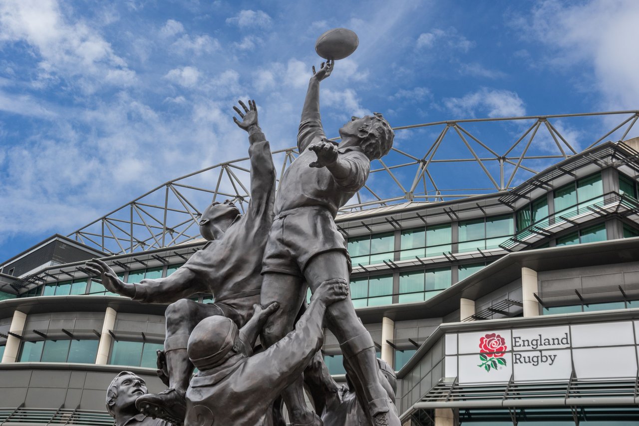 Bronze rugby lineout statue outside Twickenham Stadium, featuring the England Rugby logo on the building under a blue sky.