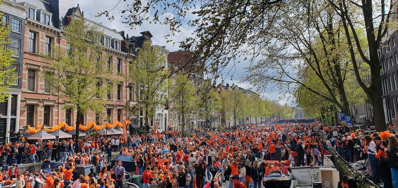 Wide shot of a bustling King's Day celebration in Amsterdam, showing thousands of people dressed in orange along a canal lined with historic buildings and trees.