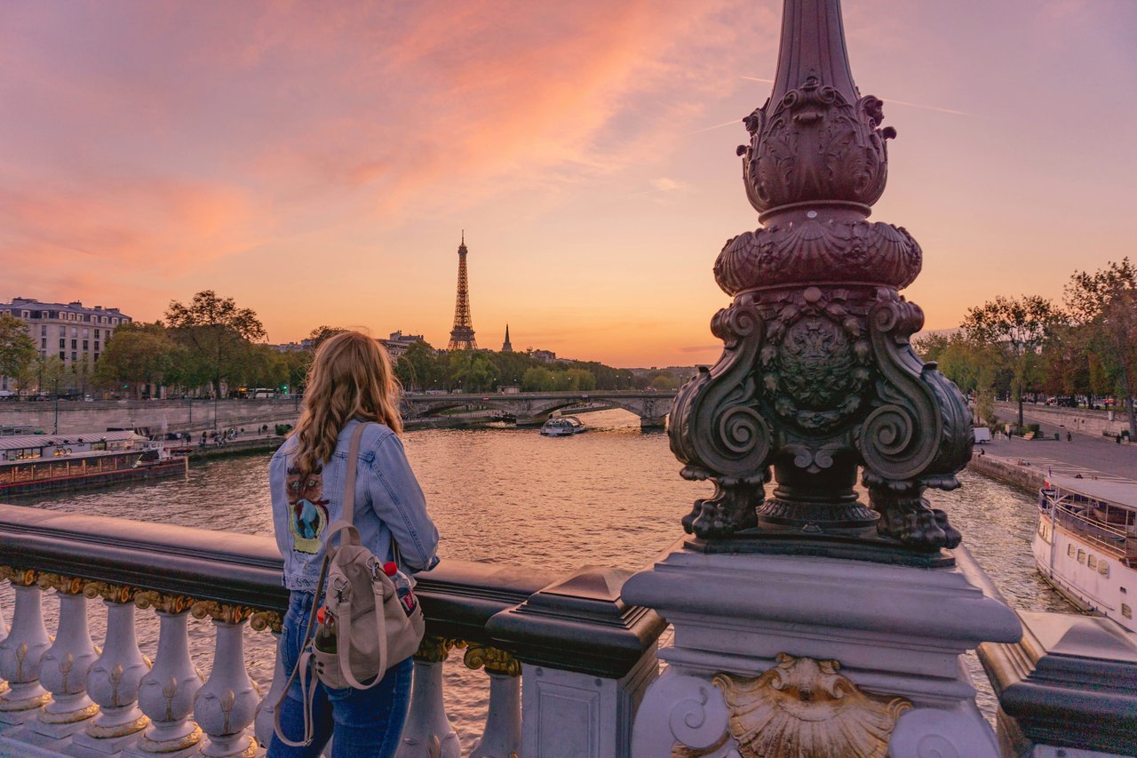 A woman with a backpack on an ornate bridge admiring the Eiffel Tower and Seine River at sunset in Paris, France.