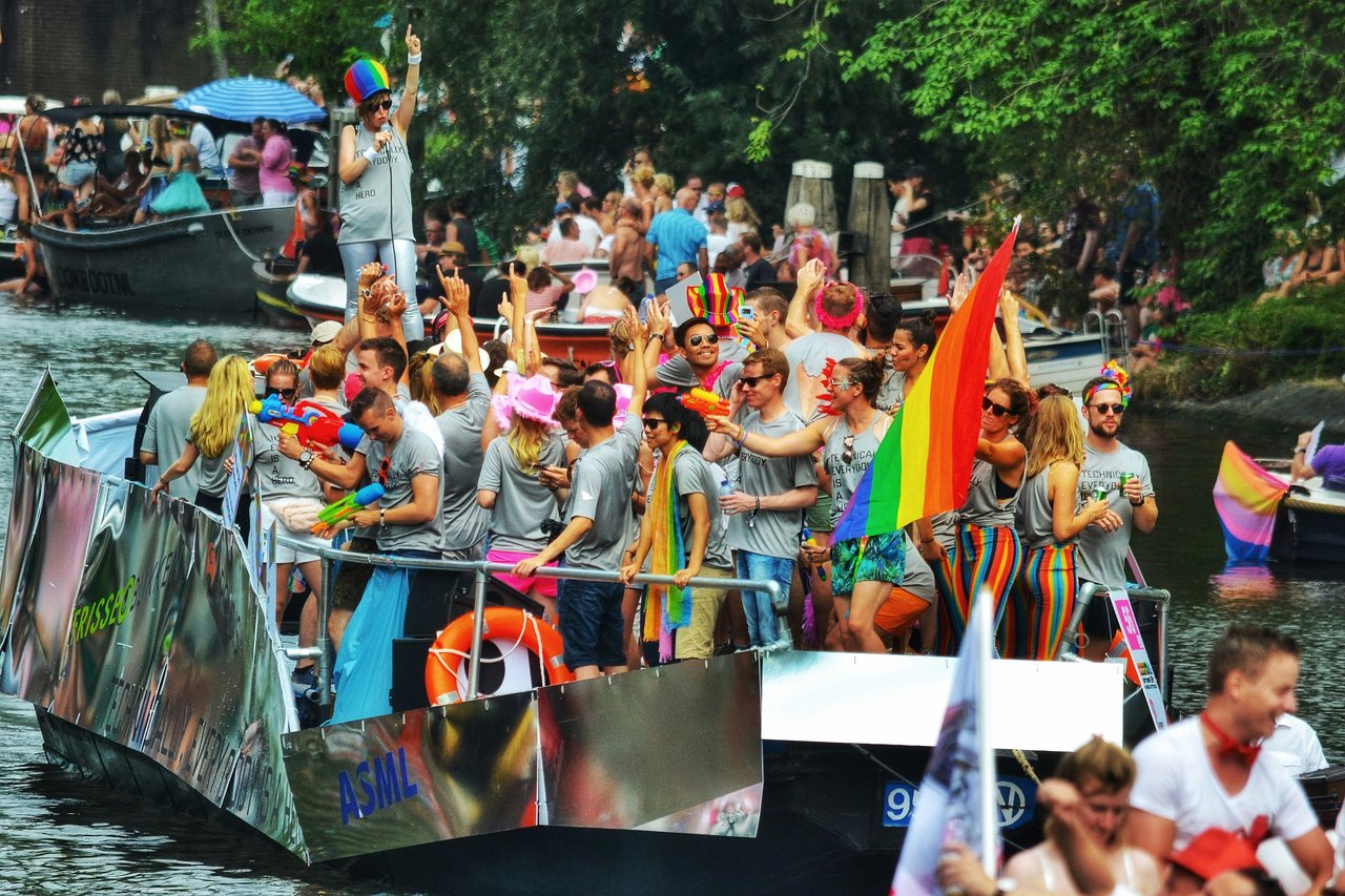 People celebrating on decorated boats in an Amsterdam canal during the Pride parade, featuring rainbow flags, colorful clothing, and water guns.