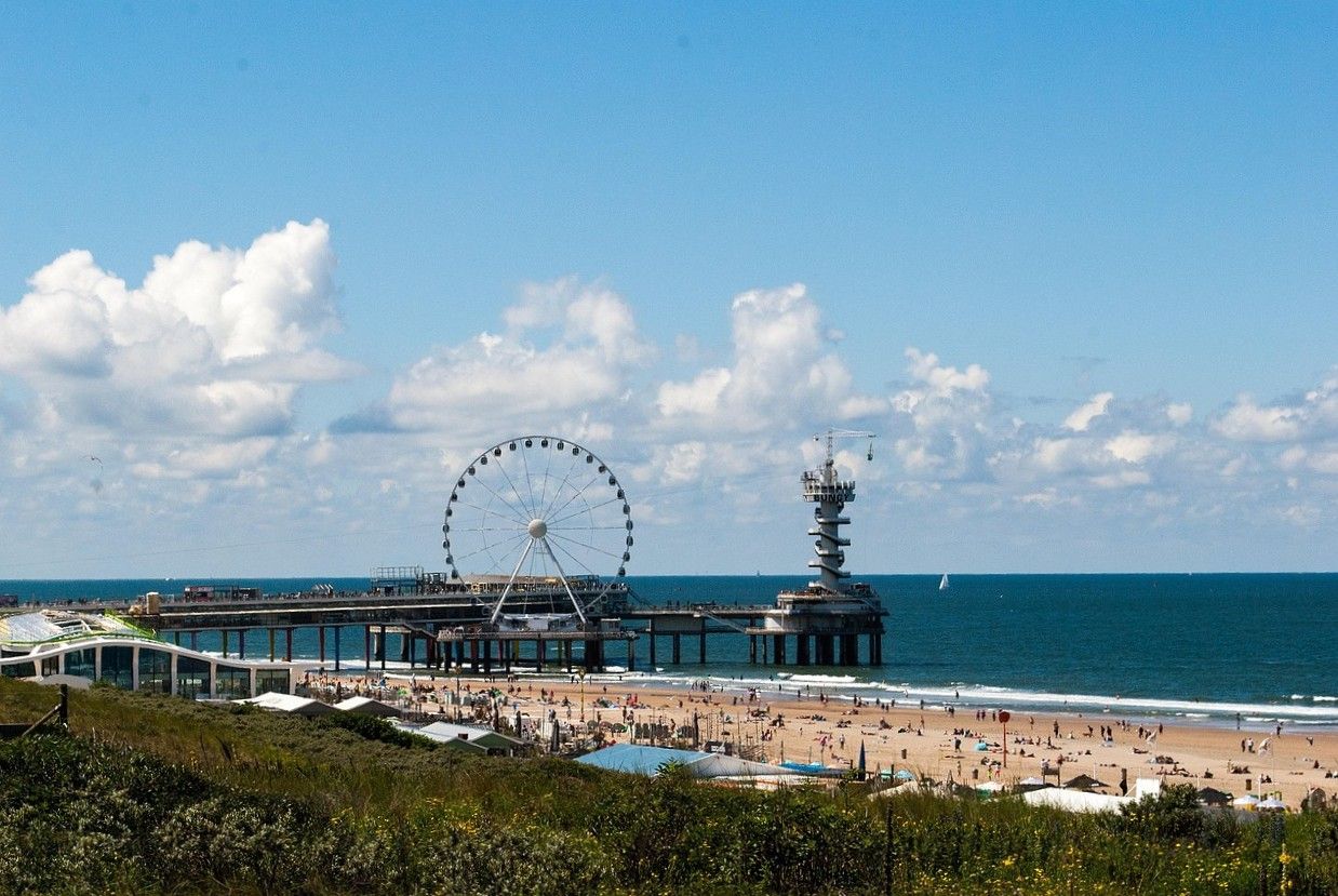 Panoramic view of Scheveningen beach and pier in The Hague, Netherlands, on a sunny day. The iconic pier features a large Ferris wheel and a bungee jumping tower, with people relaxing on the sandy shore.