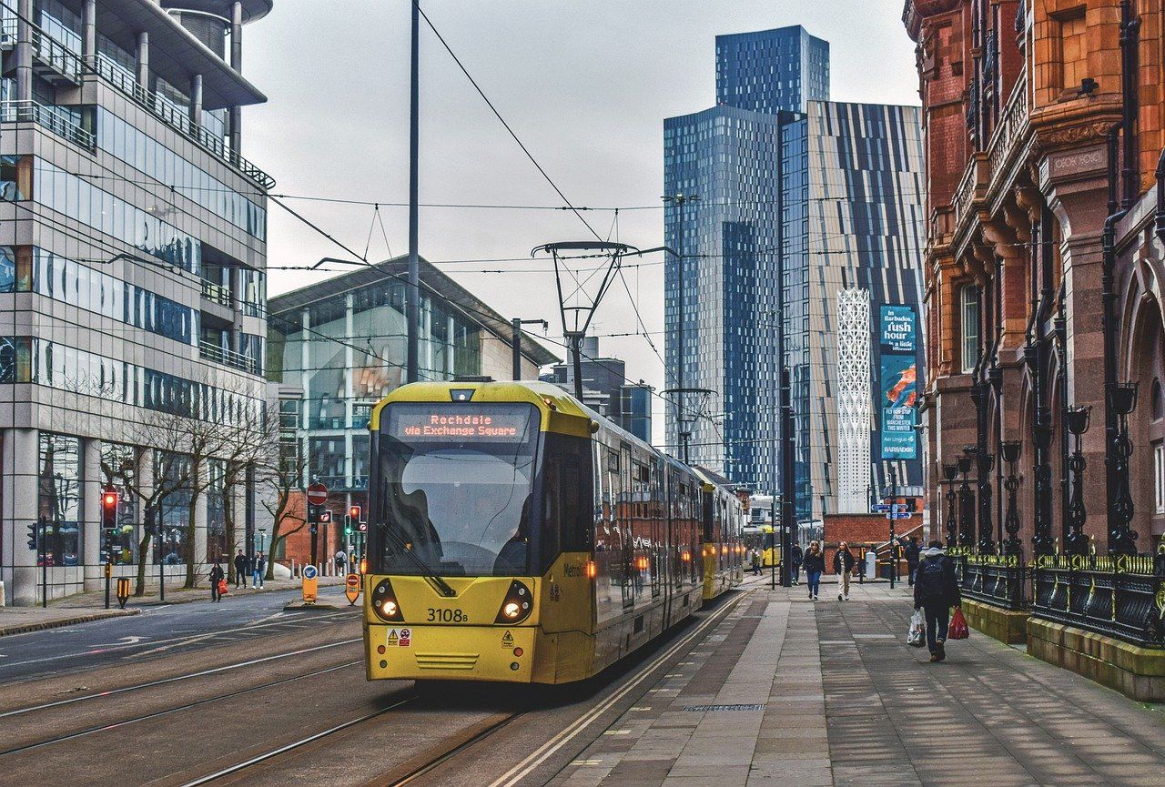 Yellow Metrolink tram travelling through Manchester city centre, flanked by modern glass skyscrapers and an ornate historic building under a cloudy sky.