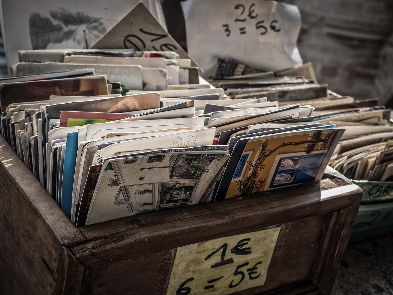 Rustic wooden crate filled with vintage postcards for sale at a European street market. Handwritten price tags show euro currency, with visible designs including architectural sketches and colorful building scenes.