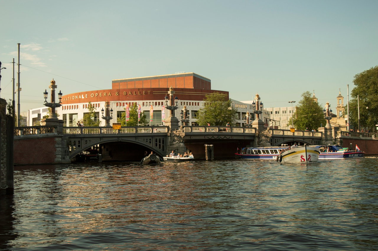 Scenic view of the Dutch National Opera & Ballet building in Amsterdam, with a classic stone bridge crossing a canal where tour boats cruise.