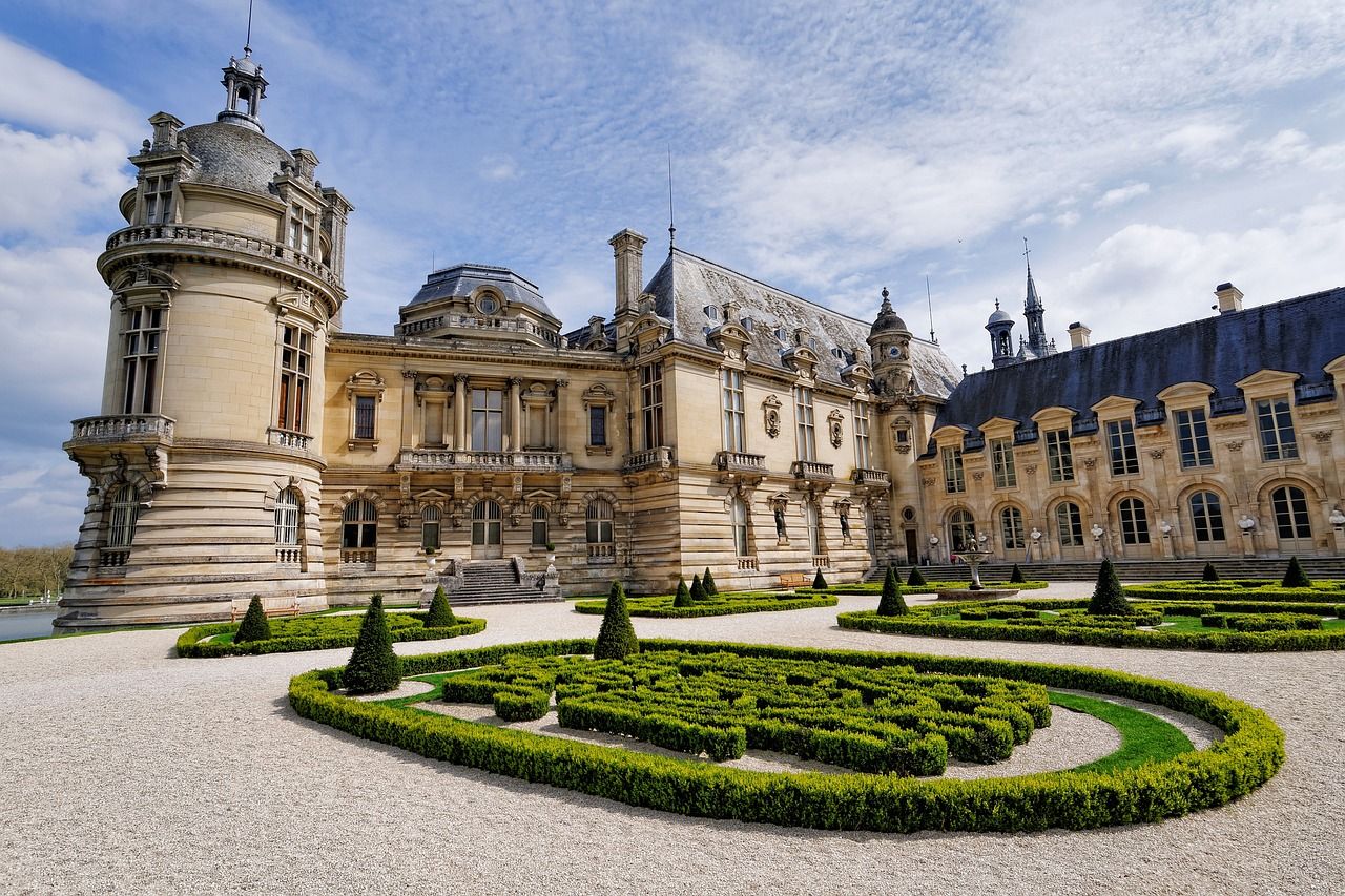 Château de Chantilly in France, a grand ornate French chateau with classical architecture, a large rounded tower, and intricate formal gardens under a partly cloudy sky.