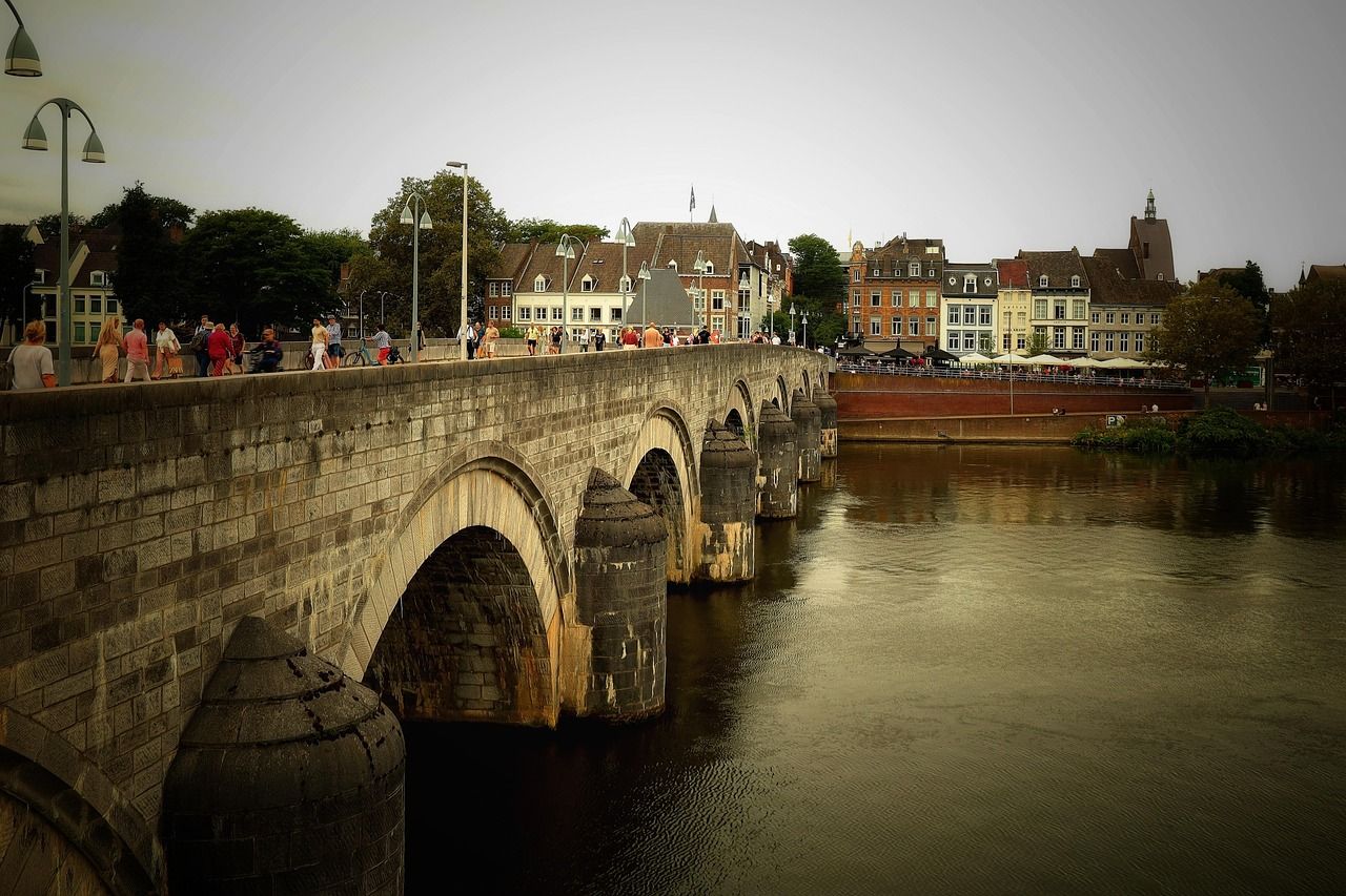 Historic Sint Servaasbrug stone bridge over the Meuse River in Maastricht, Netherlands, with pedestrians and traditional buildings lining the riverbank.