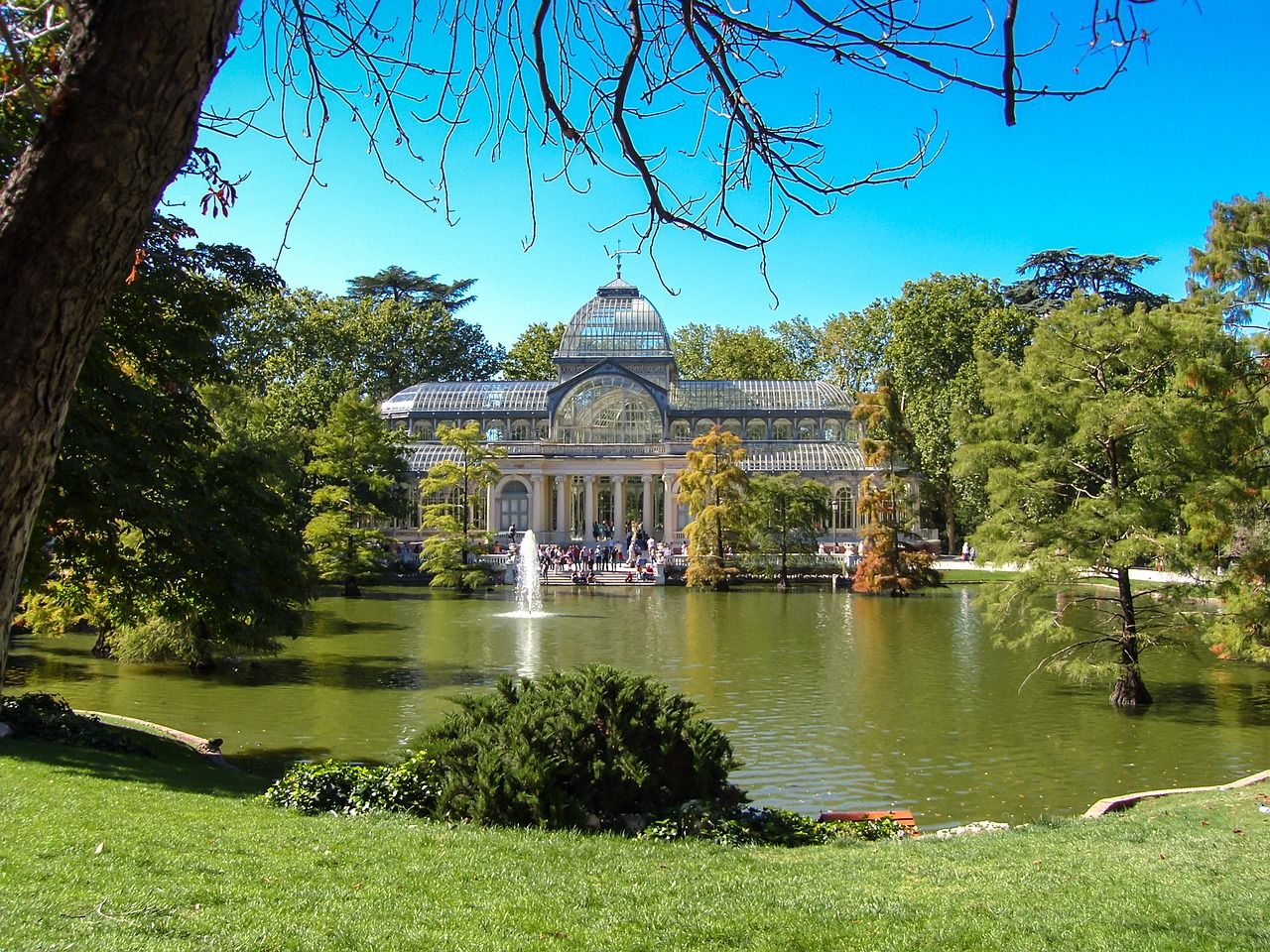 The iconic Palacio de Cristal (Crystal Palace) in Madrid's Retiro Park, a beautiful glass and iron conservatory, reflected in a large pond with a fountain and surrounded by lush green trees under a clear blue sky.