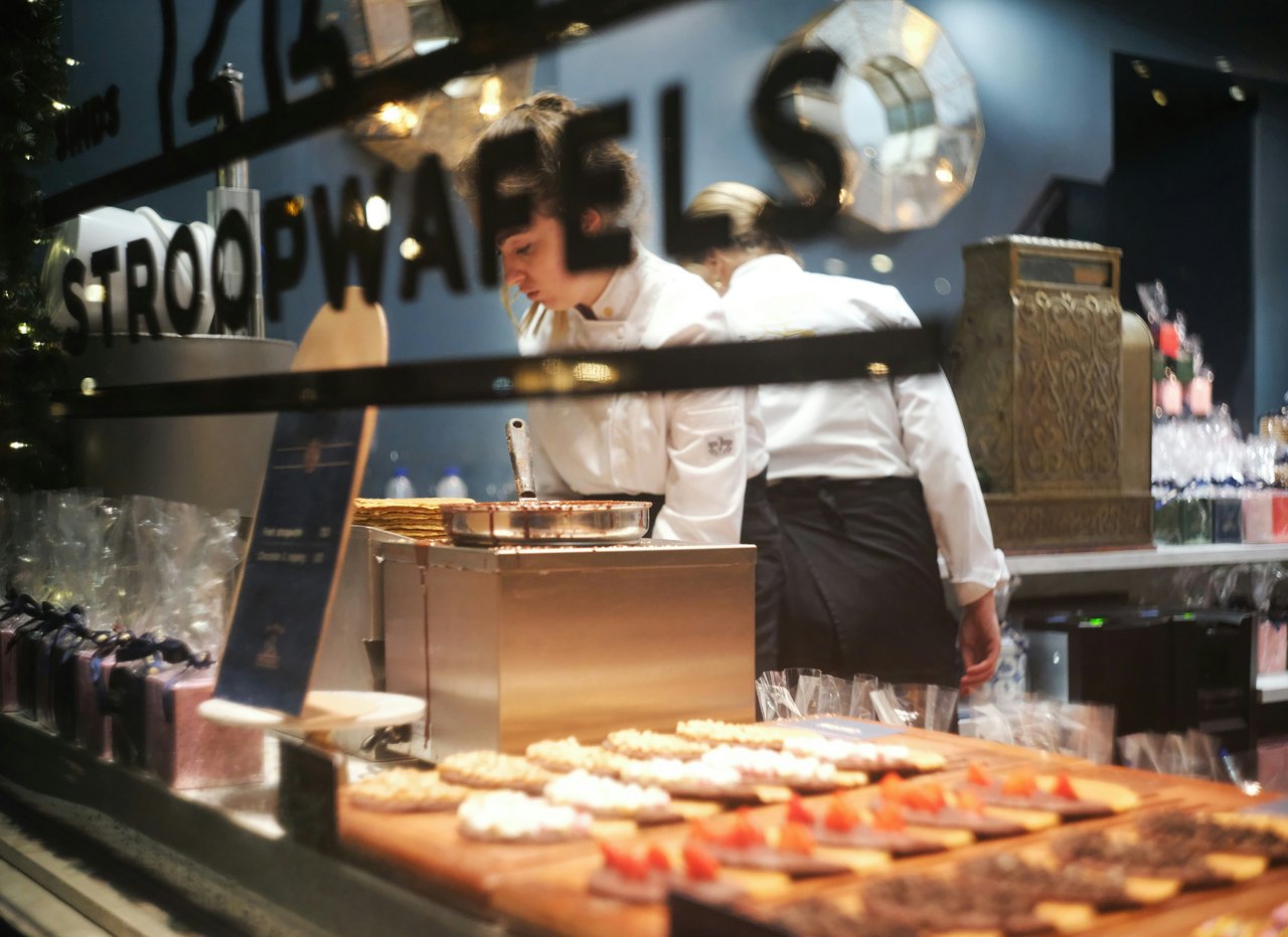 Bakers prepare fresh Dutch stroopwafels and other pastries in a shop, with a display of decorated treats in the foreground and a vintage cash register in the background.