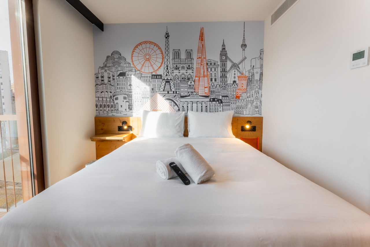 Simple double room with a bed, bedside lamp, and a wall mural depicting a cityscape of Berlin.