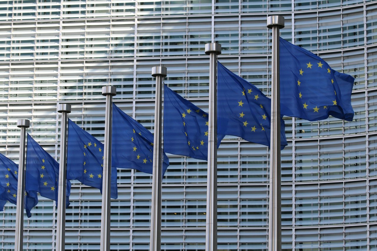 A row of blue European Union flags with gold stars waving on flagpoles in front of a modern EU institution building in Brussels, Belgium.