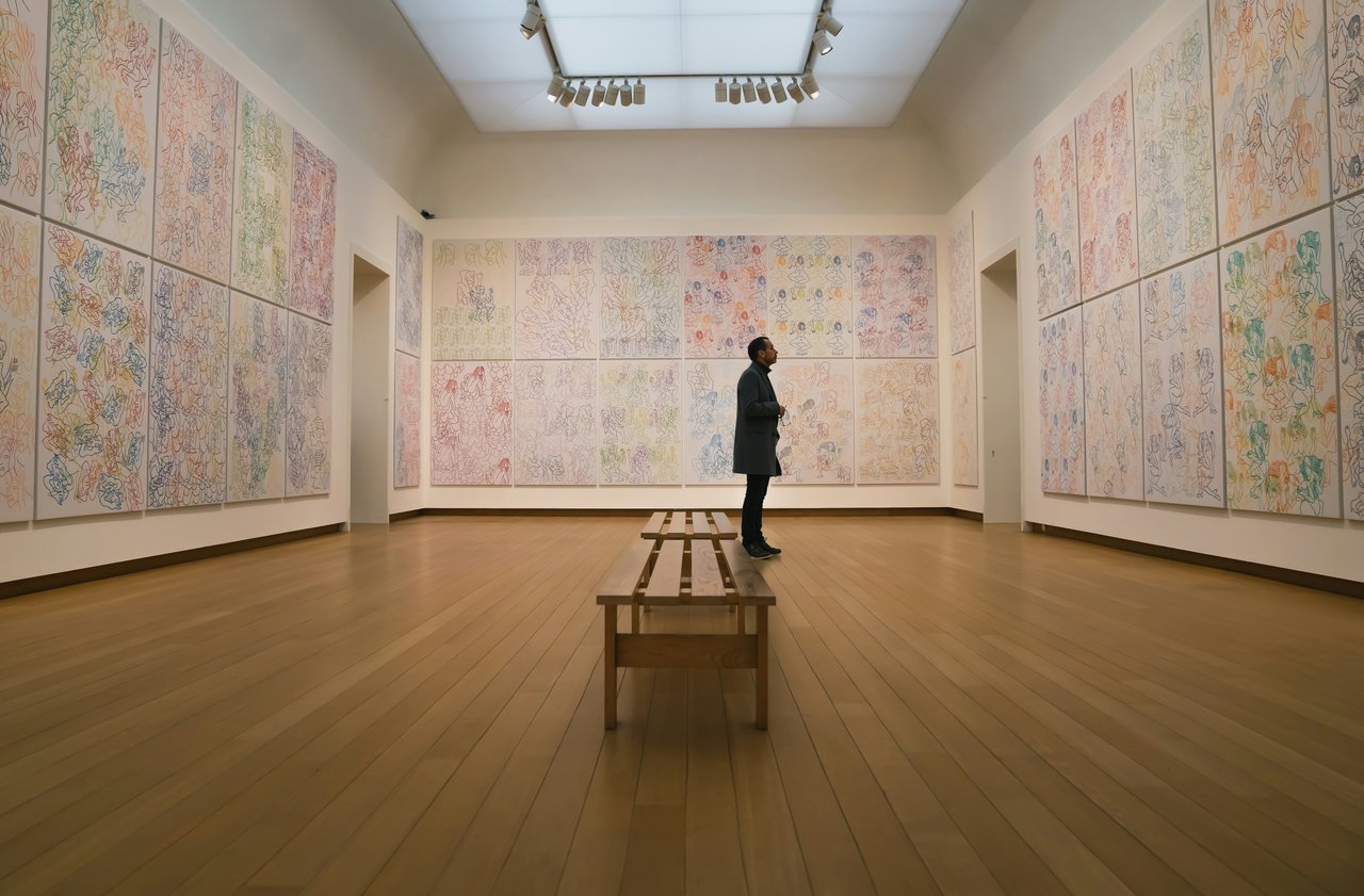 A man in a dark coat stands contemplatively in a wide, brightly lit art gallery hall, gazing at a panoramic exhibition of colorful Sol LeWitt wall drawings. The drawings feature intricate, repetitive patterns across large panels, complementing the light w
