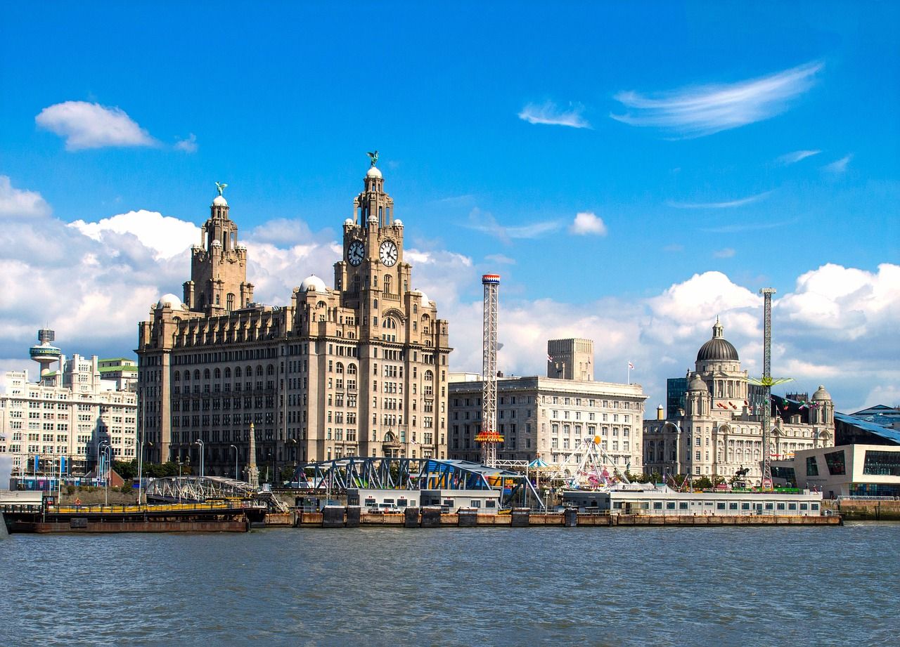 Panoramic view of Liverpool's waterfront skyline, featuring the iconic Royal Liver Building with its clock towers and Liver Birds, alongside other historic landmarks and fairground attractions on the River Mersey under a bright blue sky.