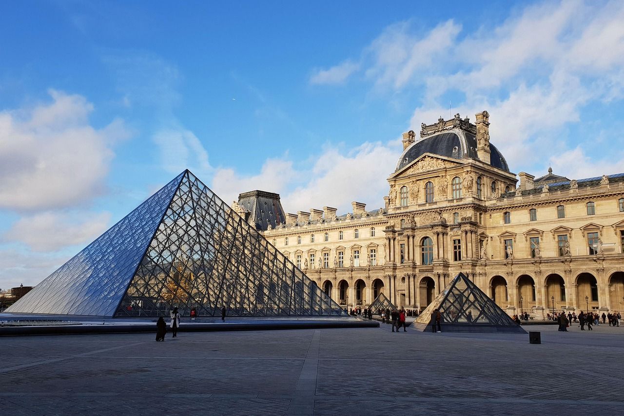 The Louvre Museum in Paris, featuring the prominent glass Louvre Pyramid on a wide paved courtyard, with the ornate classical palace building in the background under a blue, cloudy sky.