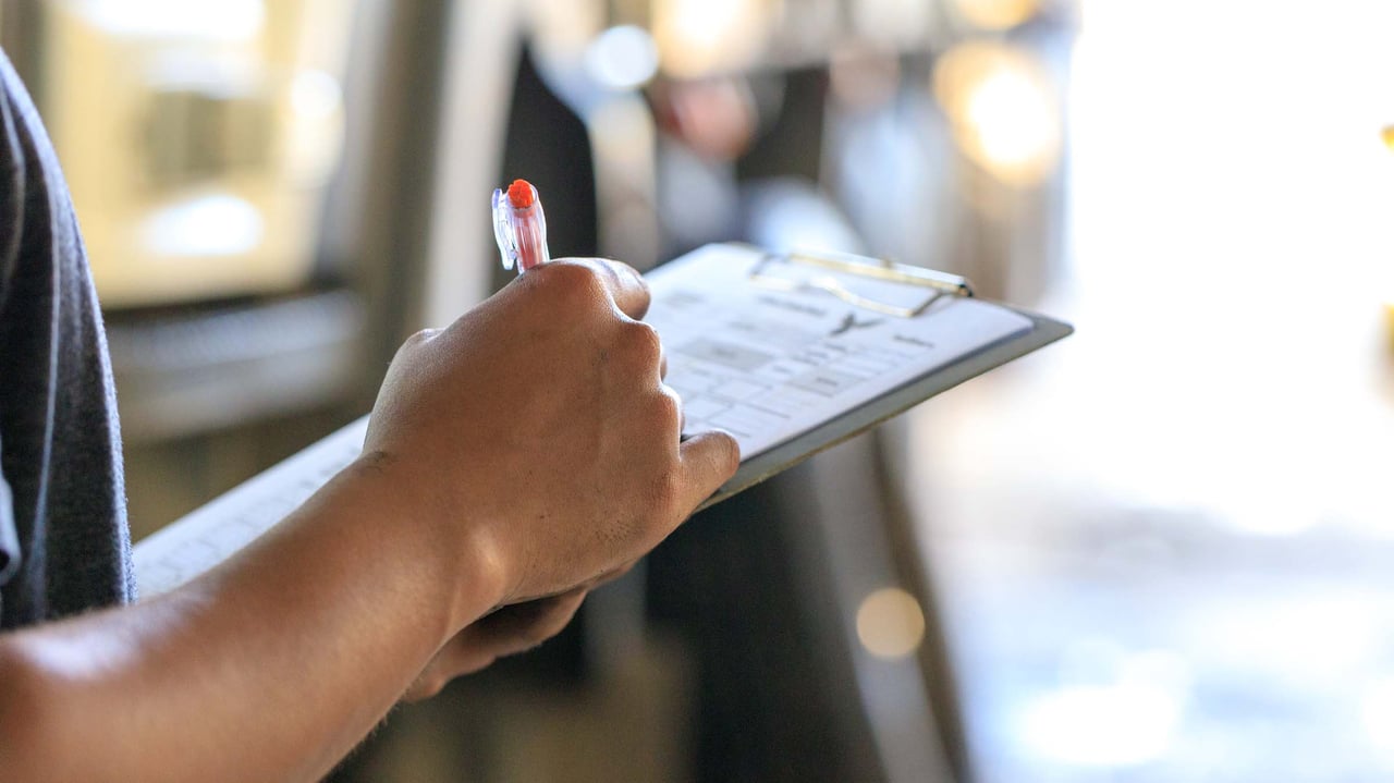 Close-up of a person's hand writing on a clipboard with a red pen, indicating data collection during a low carbon audit or sustainability assessment.