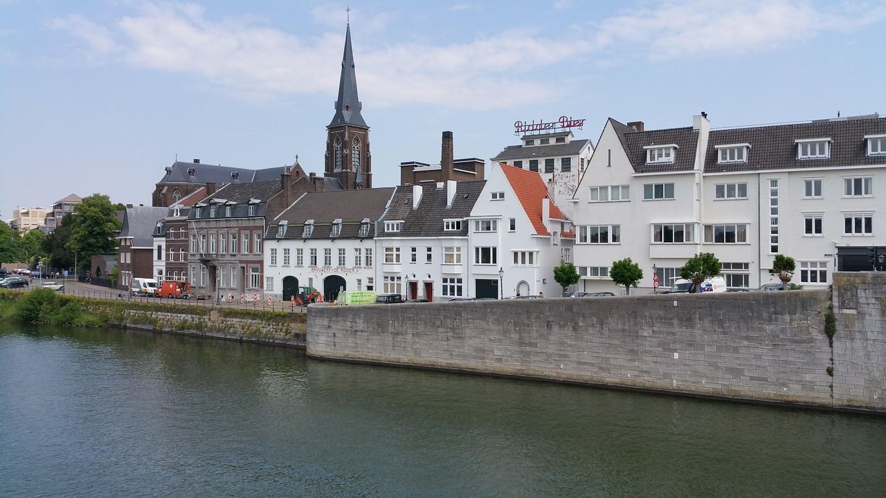 Riverside cityscape of Maastricht, Netherlands, showing historic buildings, a prominent church spire, and the Maas River in the foreground.