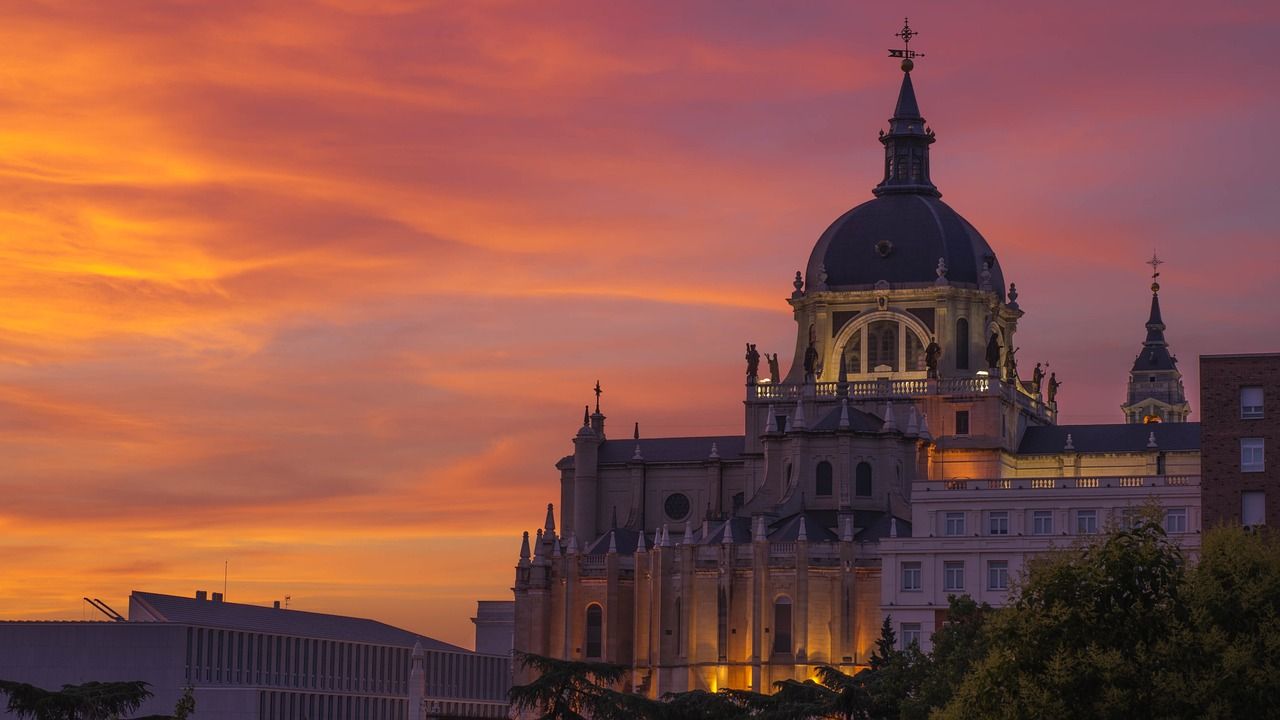 Almudena Cathedral in Madrid, Spain, illuminated at dusk against a vibrant orange and pink sunset sky.