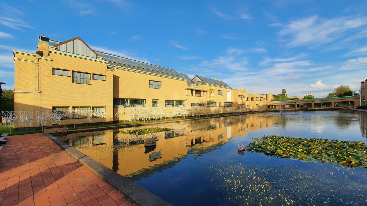 The iconic yellow brick architecture of Kunstmuseum Den Haag in The Hague, Netherlands, reflected in the calm museum pond under a blue sky.