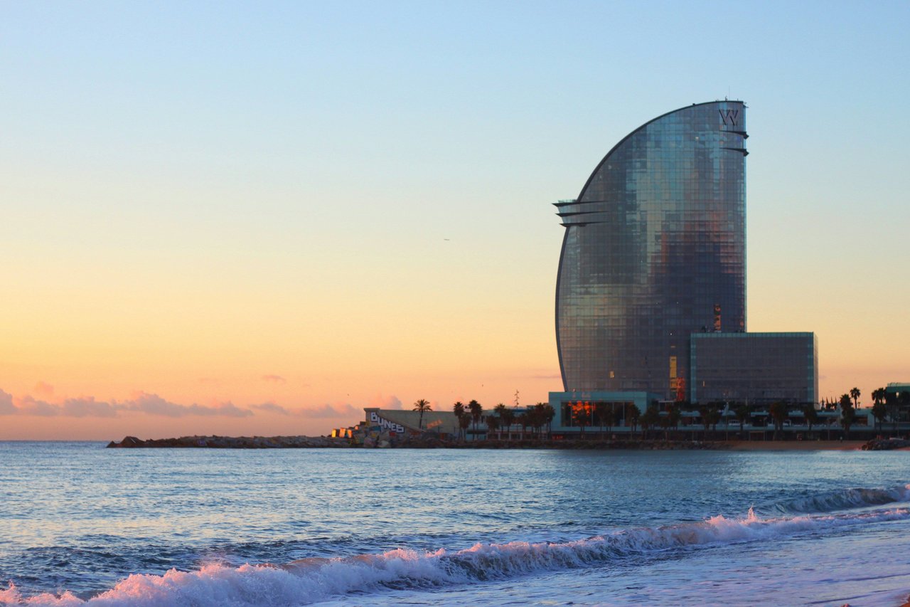 Distinctive sail-shaped hotel building in Barcelona, Spain, overlooking the Mediterranean Sea during a colorful sunrise, with waves crashing on the beach in the foreground.