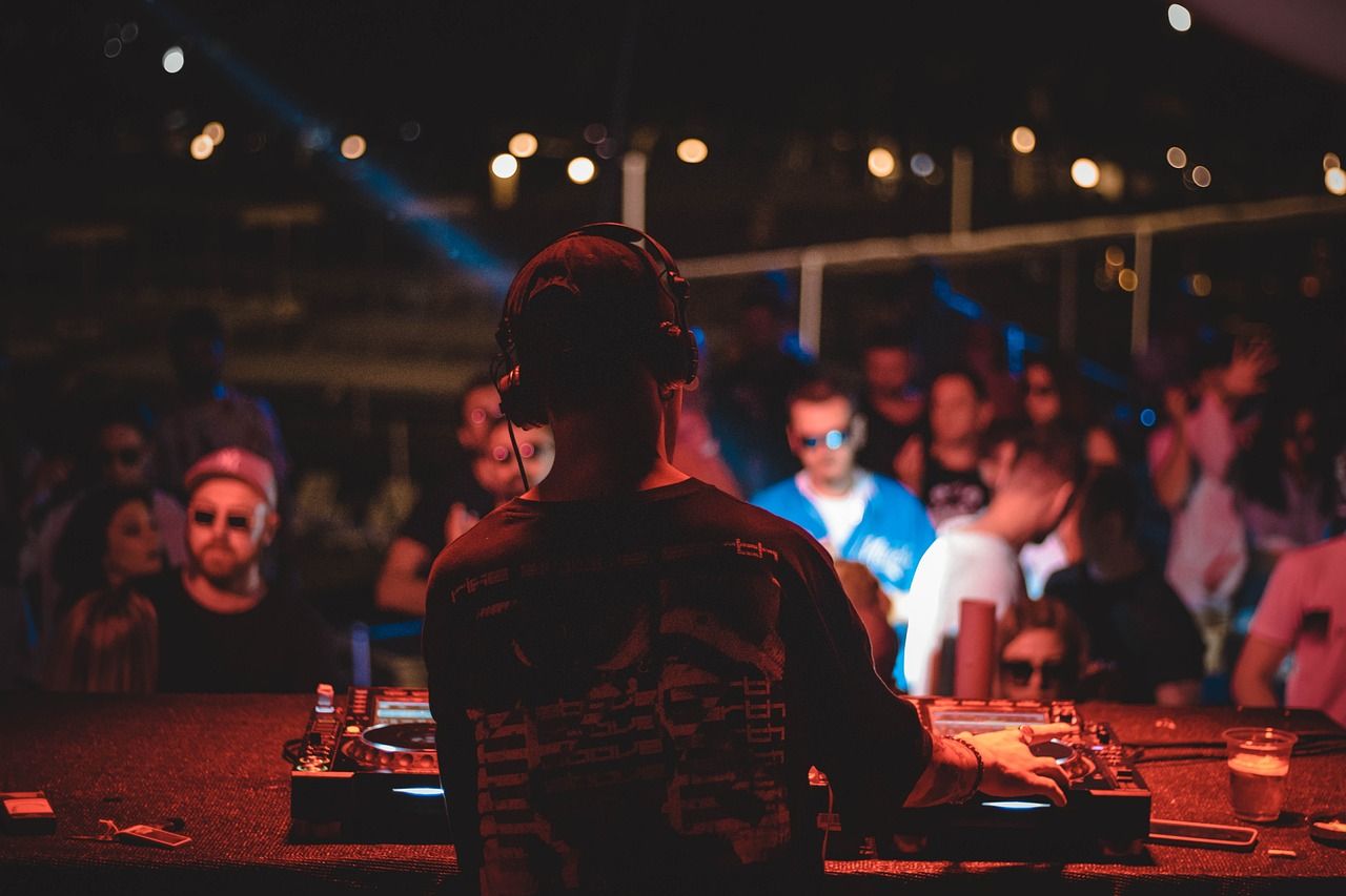 A DJ, viewed from behind, wearing headphones and actively mixing music on a console with turntables. The dark, energetic venue is lit by red and blue stage lights, with a blurred crowd enjoying the live event in the background.