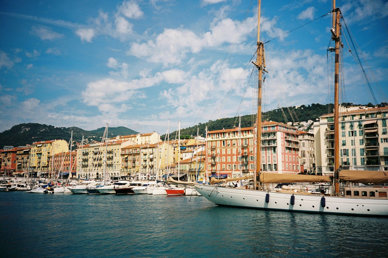 Sunny view of Port Lympia in Nice, French Riviera, with numerous sailboats and yachts docked against a backdrop of colorful pastel buildings and green hills under a blue sky.
