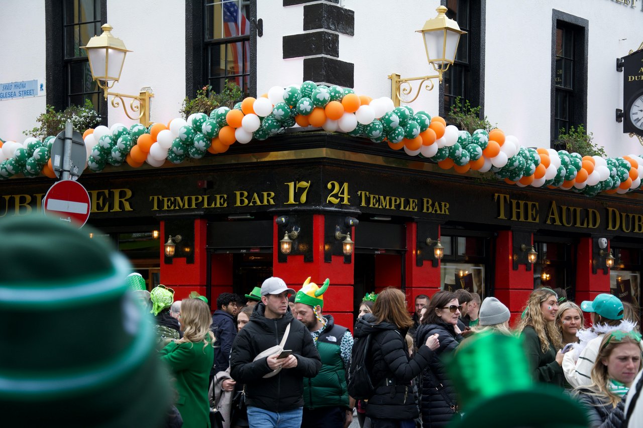 A vibrant street scene in Dublin's Temple Bar district, featuring a crowd of people celebrating St. Patrick's Day in front of a pub adorned with green, white, and orange balloons.