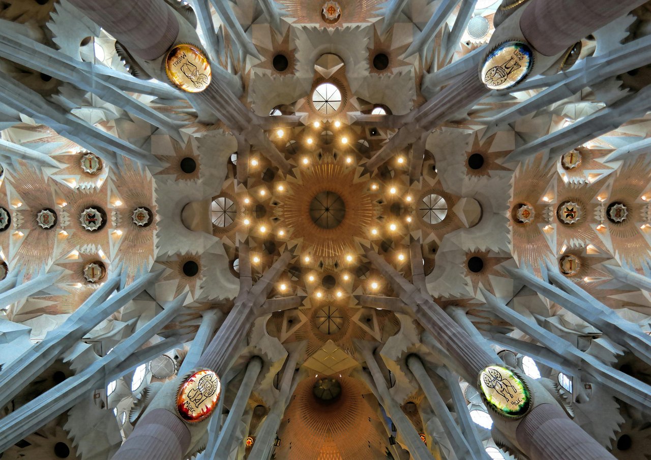 Dramatic overhead view of the Sagrada Familia basilica's ceiling in Barcelona, Spain, with towering, branching tree-like columns, intricate geometric vaults, and luminous central lighting.