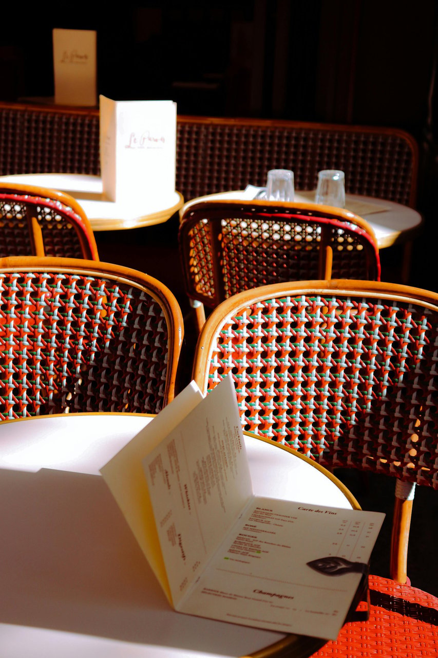 Sunny outdoor seating at a classic Parisian cafe, featuring rows of traditional wicker bistro chairs with colorful red, green, and white patterns, paired with small round white tables. An open menu displaying "Carte des Vins" and other French text rests o