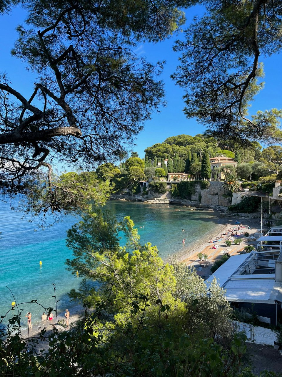 Beach scene with turquoise water, a sandy shore, and buildings nestled on a hillside, framed by trees.