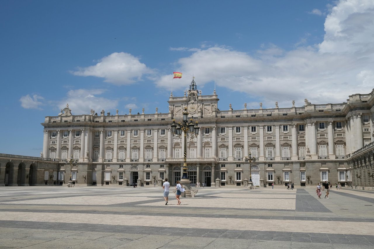 Royal Palace of Madrid, a grand historic building in Spain, viewed from the expansive Plaza de la Armería on a sunny day with a Spanish flag flying.