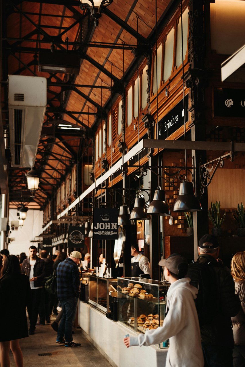 A bustling indoor gourmet food market in Madrid, Spain, featuring a high arched ceiling with exposed brick and ironwork, ornate wooden stalls, diverse food vendors, and people browsing; visible signs include 'Brioches' and 'John T Barrita'.