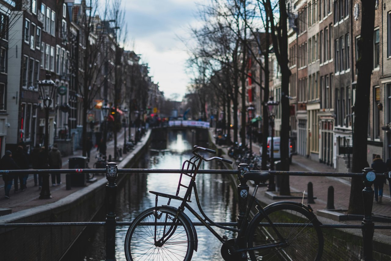 Iconic view of an Amsterdam canal with a black bicycle parked on a bridge railing, flanked by historic gabled buildings and bare trees under a cloudy sky.