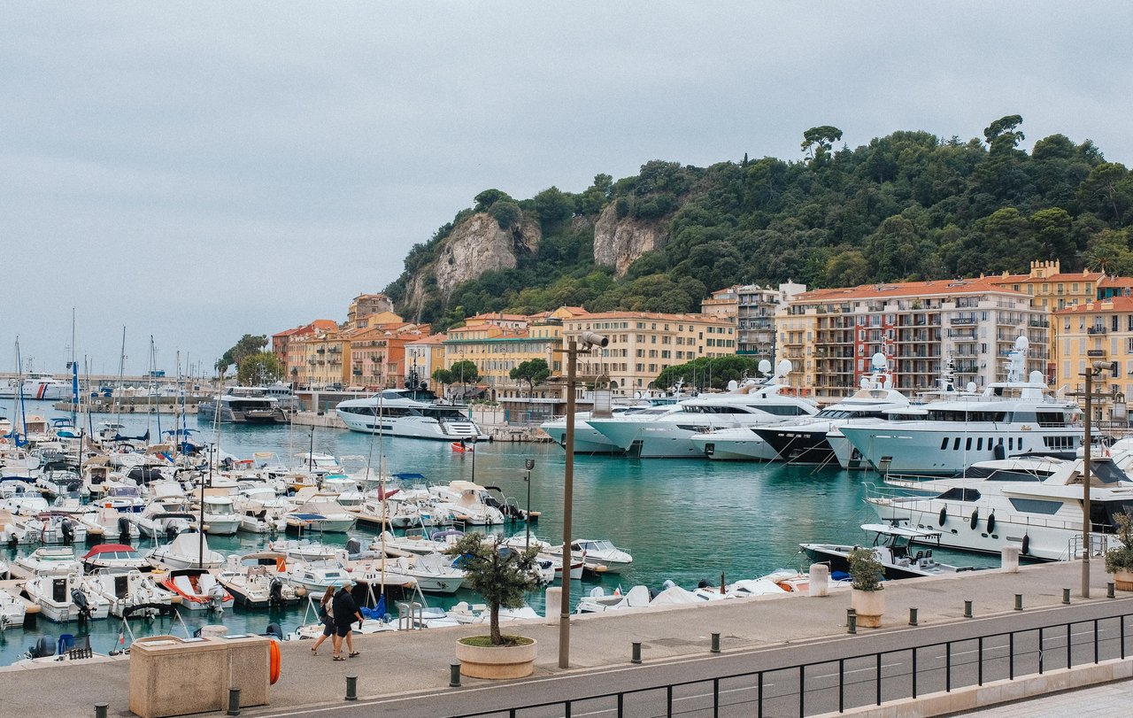Scenic view of Port Lympia in Nice, France, showcasing luxury yachts and numerous boats docked in the vibrant harbor with colorful waterfront buildings and a lush green hill in the background.
